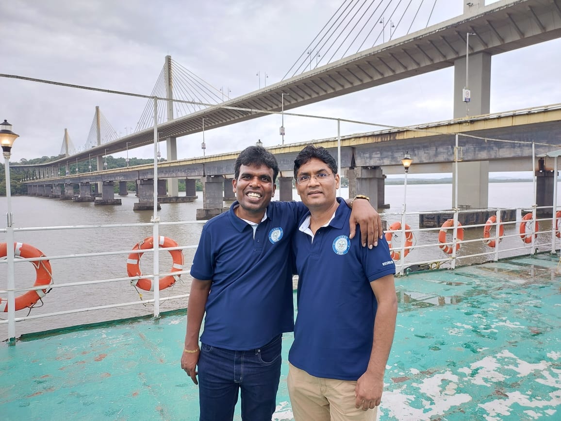 Two DRDO friends on a boat with a bridge behind them