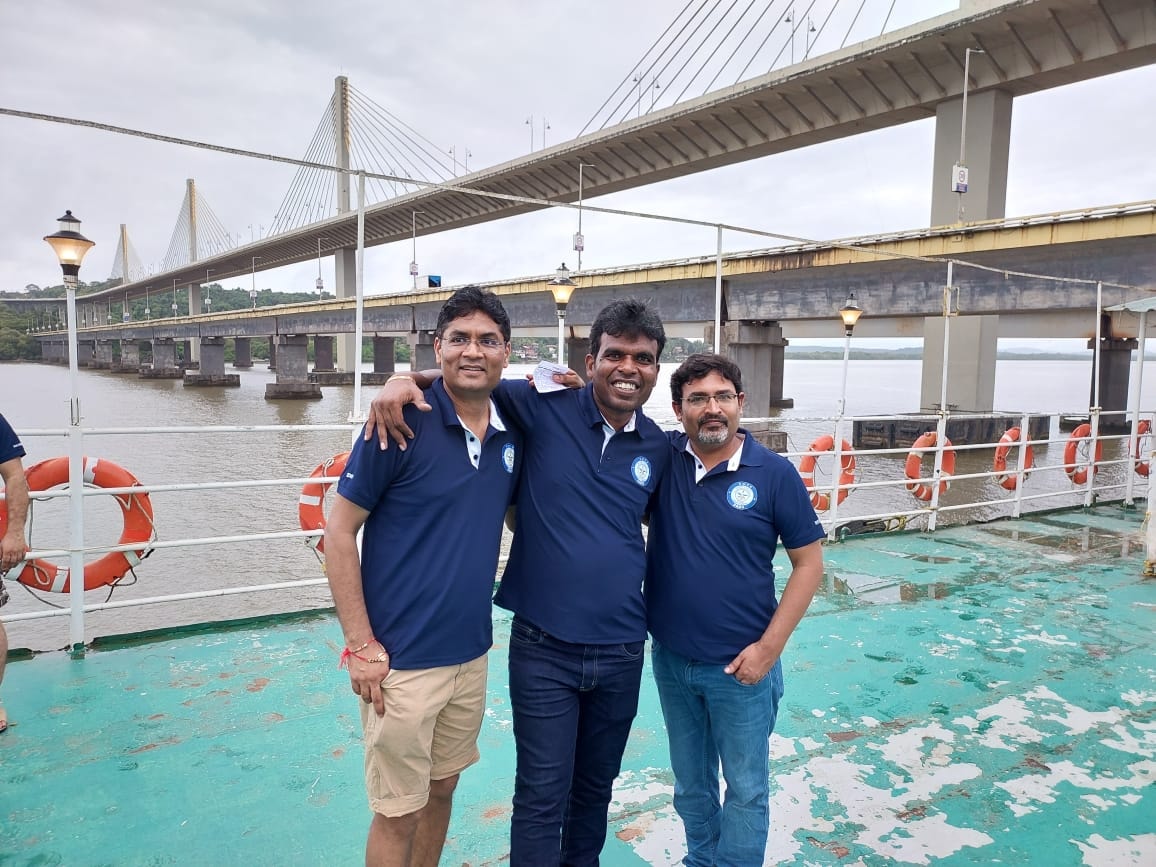Three DRDO friends on a boat with a waterfront bridge behind them